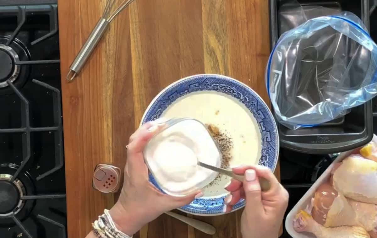 A person is seen from above preparing to mix ingredients in a bowl on a wooden countertop. The bowl contains a liquid and spices. The person is holding a small container of a white, creamy substance and a spoon. Chicken pieces and kitchen tools are nearby.