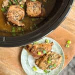 A top view of a slow-cooker with cooked chicken pieces garnished with chopped green onions. Below the slow cooker, there is a plate on a wooden surface with two pieces of chicken also garnished with green onions.