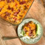 A glass baking dish filled with cheesy, bacon-topped casserole sits on a wooden table. Next to it, a serving of the casserole is on a patterned plate with a fork and some lettuce.