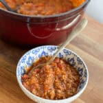 A bowl of chunky tomato-based sauce with ground meat is placed on a wooden surface. The sauce is served in a white bowl with blue decorative patterns on the inside. A spoon rests in the bowl. In the background is a red pot filled with more of the sauce.