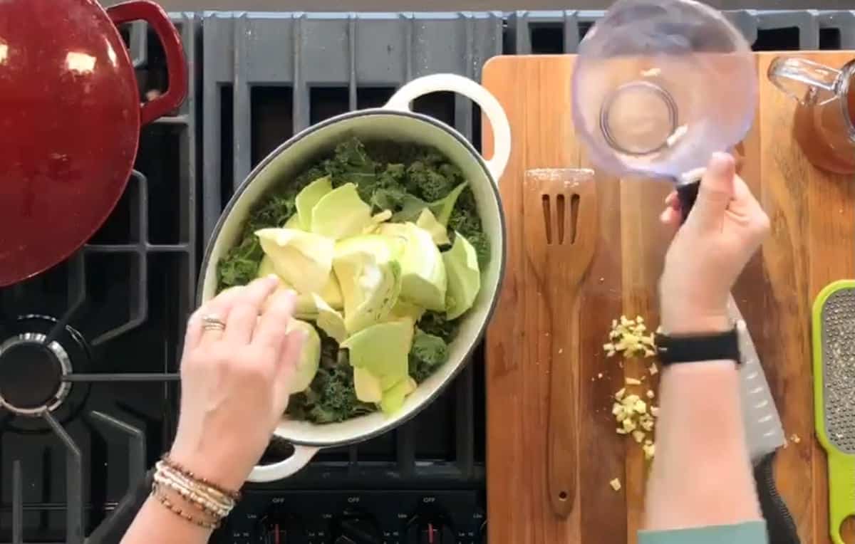 A person is preparing a meal on a stovetop. They are adding sliced cabbage to a pot already filled with leafy greens. The kitchen counter has a cutting board with minced garlic and a knife, and there's a red pot nearby.