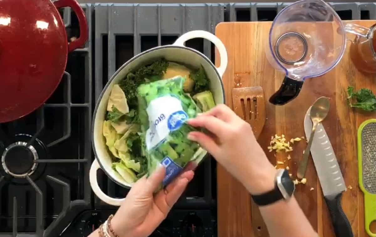 Overhead view of a kitchen scene. A person is adding a bag of chopped green vegetables into a white cooking pot on a stove. Nearby are chopped ingredients, a knife, a spatula, a grater, and a blender on a wooden cutting board. A red pot is on the stove.