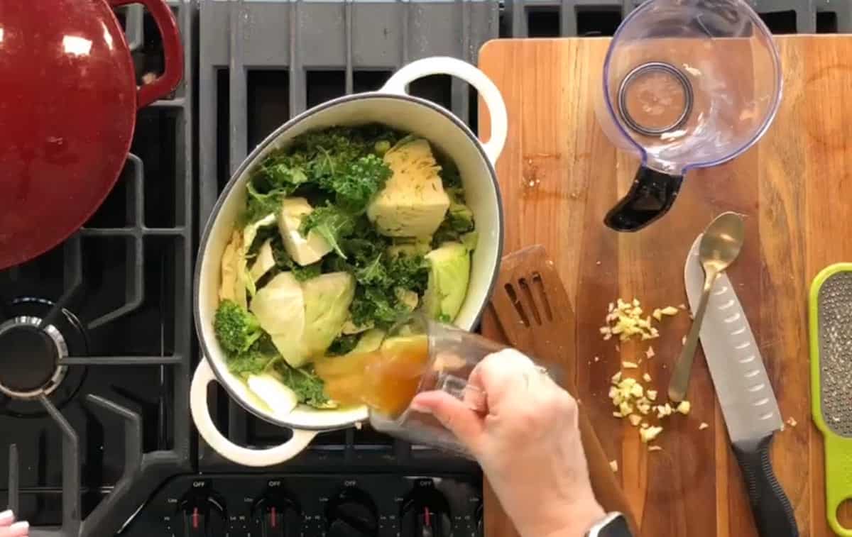 Overhead view of a person pouring liquid into a pot containing chopped vegetables on a stove. A wooden cutting board with knives, chopped garlic, a grater, and an empty measuring cup are placed beside the stove. A red pot is on the left burner.