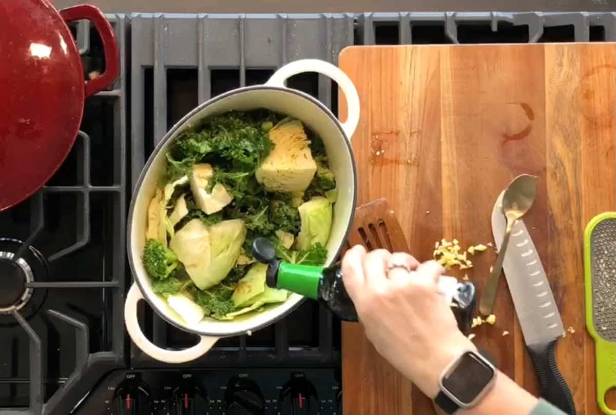 A person is pouring a dark liquid, likely oil, into a pot filled with leafy greens and chopped vegetables on a stovetop. Nearby, various kitchen utensils and chopped garlic rest on a wooden cutting board. A red pot sits next to the stove burner.