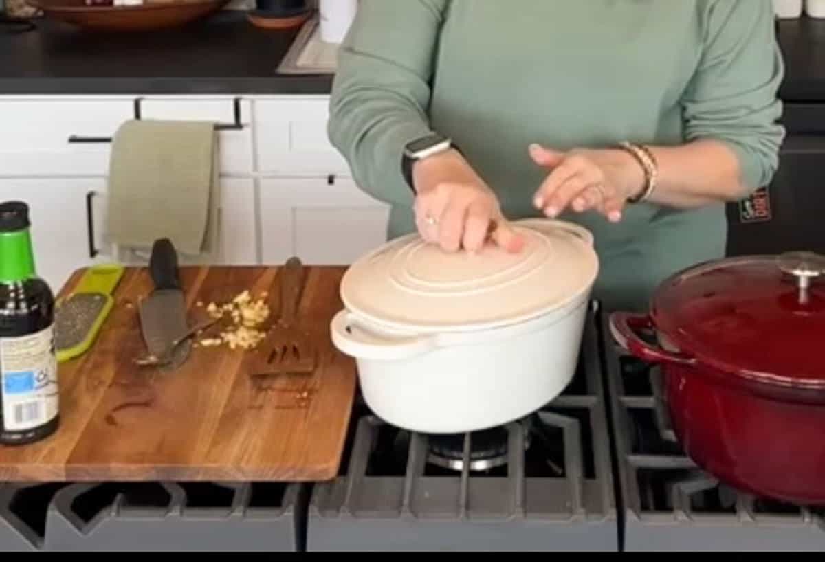 Person in a green top stands in a kitchen, placing a lid on a white pot on a stove. A red pot sits next to it. A cutting board with a knife, chopped garlic, and seasonings is on the counter nearby. A green towel hangs from a drawer.