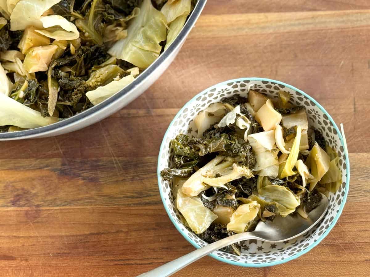 A bowl of cooked greens, including kale and cabbage, is placed on a wooden surface. A spoon is resting in the bowl. There is also a larger metallic bowl filled with more of the greens in the background.