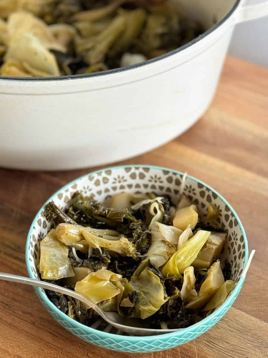 A bowl of cooked leafy green vegetables, including cabbage, placed on a wooden surface with a fork in the bowl. A larger white pot in the background contains more of the same cooked vegetables.