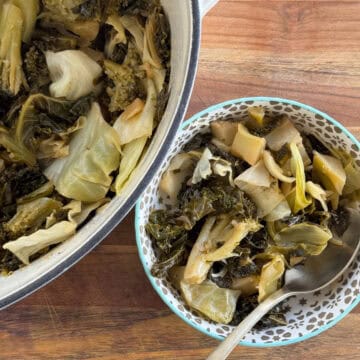 A bowl of cooked collard greens with a spoon placed beside a larger pot containing more of the same dish. The greens are wilted and mixed, sitting on a wooden surface. The smaller bowl has a decorative pattern on its rim.