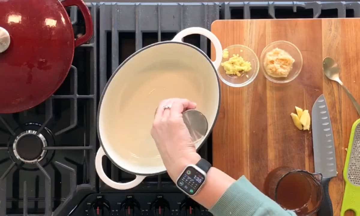 A person is seen pouring liquid from a small measuring cup into a white oval pot on a stovetop. Nearby, a cutting board holds a knife, a grater, and three small bowls containing minced ingredients. A red pot is also visible on the stove. The person wears a smartwatch.