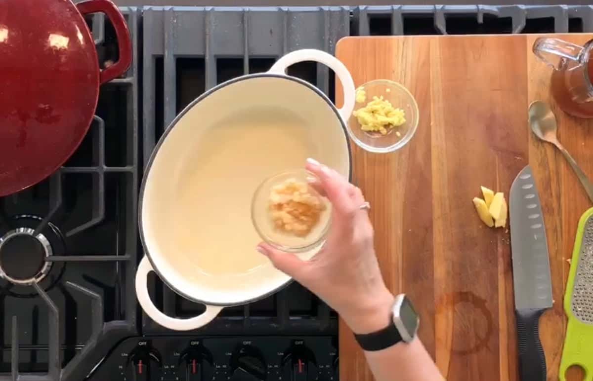 Top-down view of a kitchen counter. A person's hand is adding minced garlic from a small bowl into a white pot on a stove. Nearby, there is a cutting board with some chopped ginger, a knife, a spoon, and a grater. A red pot sits on another burner.