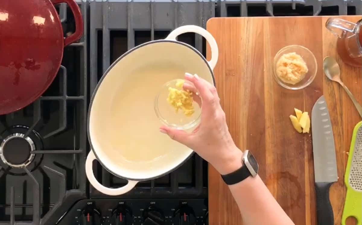 A person wearing a smartwatch is adding minced garlic from a small bowl into a white, oval-shaped Dutch oven placed on a stove. There are other bowls, a knife, and a wooden cutting board on the countertop beside the stove.