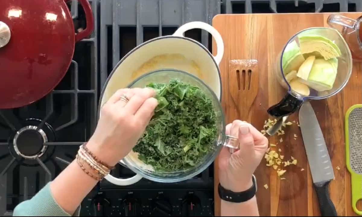 A person is adding kale from a glass bowl into a white pot on a stove. To the right, a wooden cutting board shows chopped garlic, an avocado in a jar, a knife, and a food grater. A red pot sits to the left on another burner.