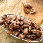 A glass dish filled with candied pecans sits on a wooden surface. Several pecans are scattered nearby on the wood. The pecans are coated in a brown, crystalline sugary layer.