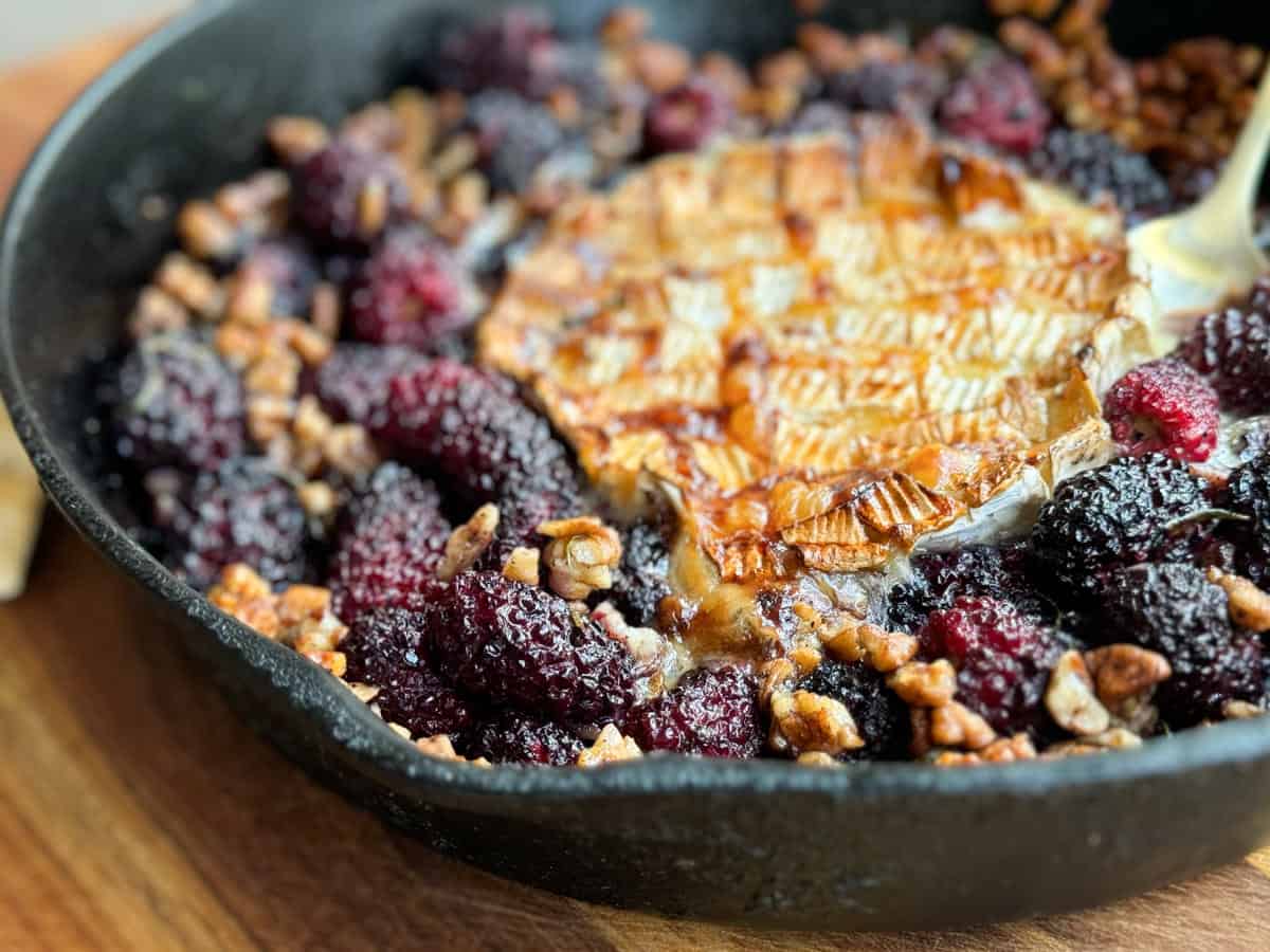 A cast iron skillet containing a baked Brie cheese wheel, surrounded by blackberries and pecans. The cheese is golden-brown and slightly melted, with a serving utensil partially visible on the right. The skillet is placed on a wooden surface.