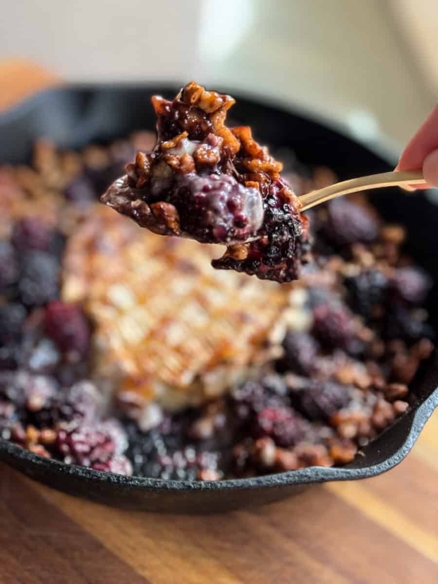 A metallic spoon holds a bite of blackberry crisp with a crunchy topping, above a cast-iron skillet filled with the dessert. The background is slightly blurred, highlighting the focused view of the spoonful.