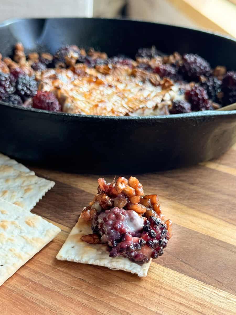 A close-up image shows a piece of matzo cracker topped with a mixture of nuts and blackberries placed in front of a cast iron skillet containing more of the nut and blackberry mixture. The scene is set on a wooden surface.