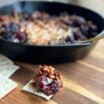 A black cast iron skillet contains a baked dish with mixed blackberries and a caramelized topping. A piece of this dish is spooned onto a cracker resting on a wooden cutting board in the foreground.