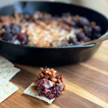 A black cast iron skillet contains a baked dish with mixed blackberries and a caramelized topping. A piece of this dish is spooned onto a cracker resting on a wooden cutting board in the foreground.