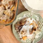 A top-view image of a serving of bread pudding on a decorative plate next to a glass baking dish filled with more bread pudding. The serving on the plate is topped with a creamy sauce, and a fork is placed beside it.