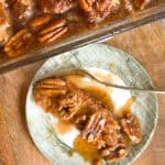 A plate with a slice of pecan pie topped with whole pecans and a rich caramel-like sauce. A fork rests on the plate, and the remaining pecan pie is visible in a baking dish in the background. The table surface is wooden.