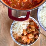 A bowl of gumbo with shrimp, okra, and other vegetables in a tomato-based sauce is served next to a pot of the same gumbo. The bowl is half-filled with white rice. Another bowl of white rice is partially visible on the right side.