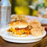 A cheeseburger with melted cheese, pickles, ketchup, and a toasted bun is on a white plate next to a serving of chips. A can of soda is also present in the background, along with blurred elements of food on a wooden table.