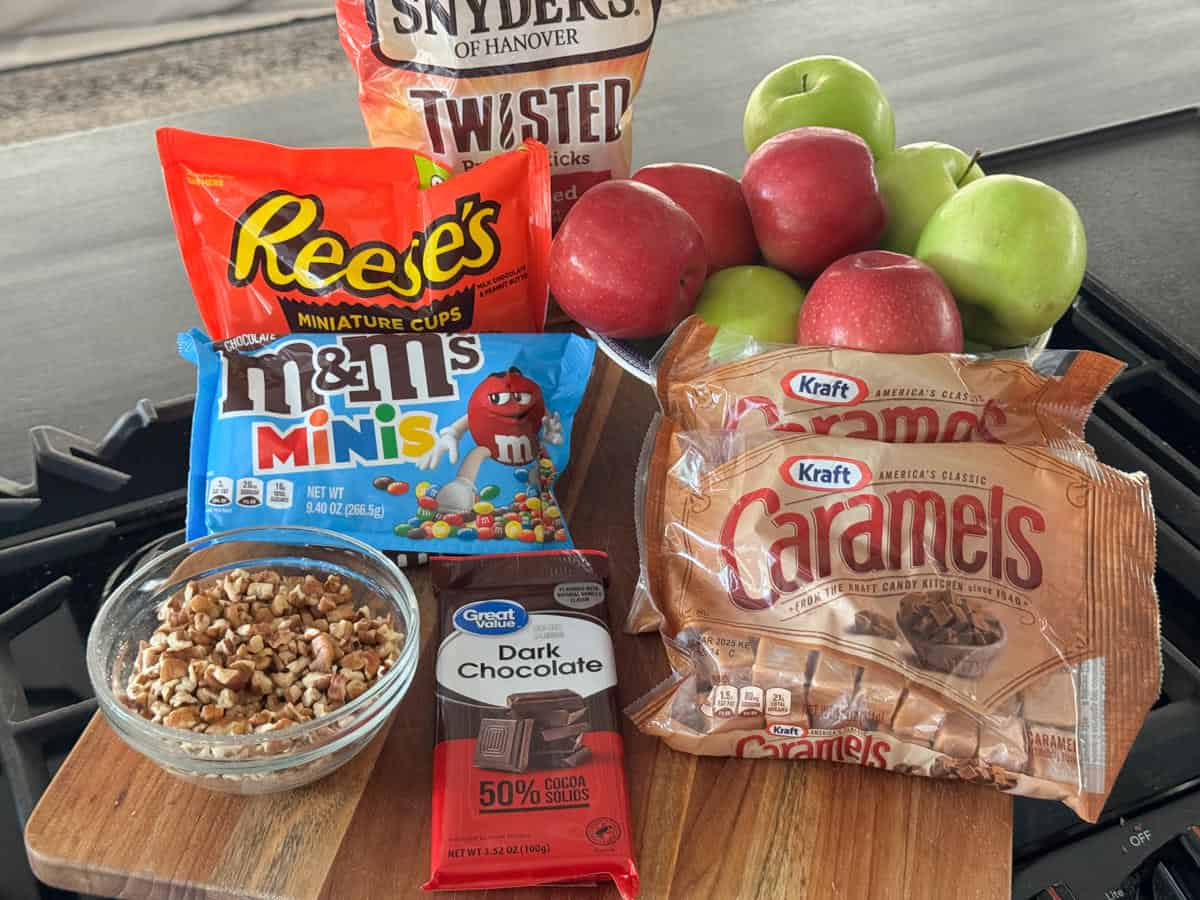 A display of ingredients for making caramel apples, including Snyder's pretzels, Reese's miniature cups, M&M's minis, Kraft caramels, dark chocolate, a bowl of chopped nuts, and a bunch of green and red apples.