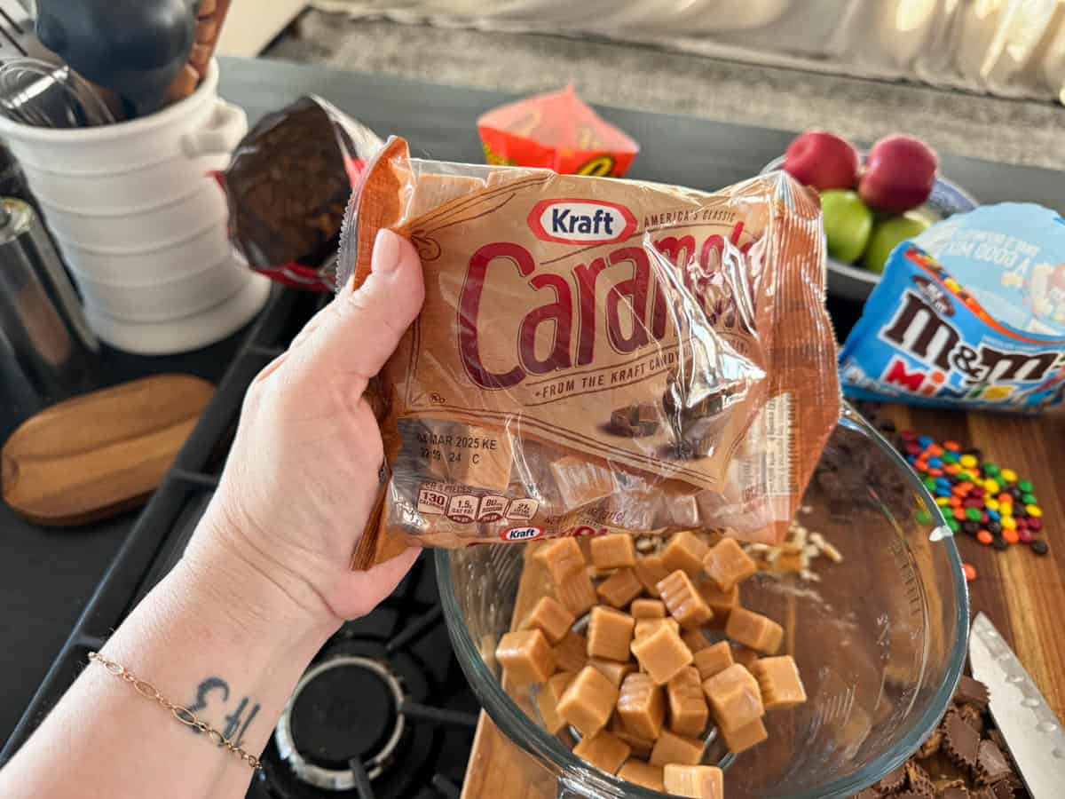 A hand holding a bag of Kraft Caramels above a bowl containing unwrapped caramels. The countertop has various ingredients and snacks including a package of M&M's, fresh apples, and a wooden cutting board.