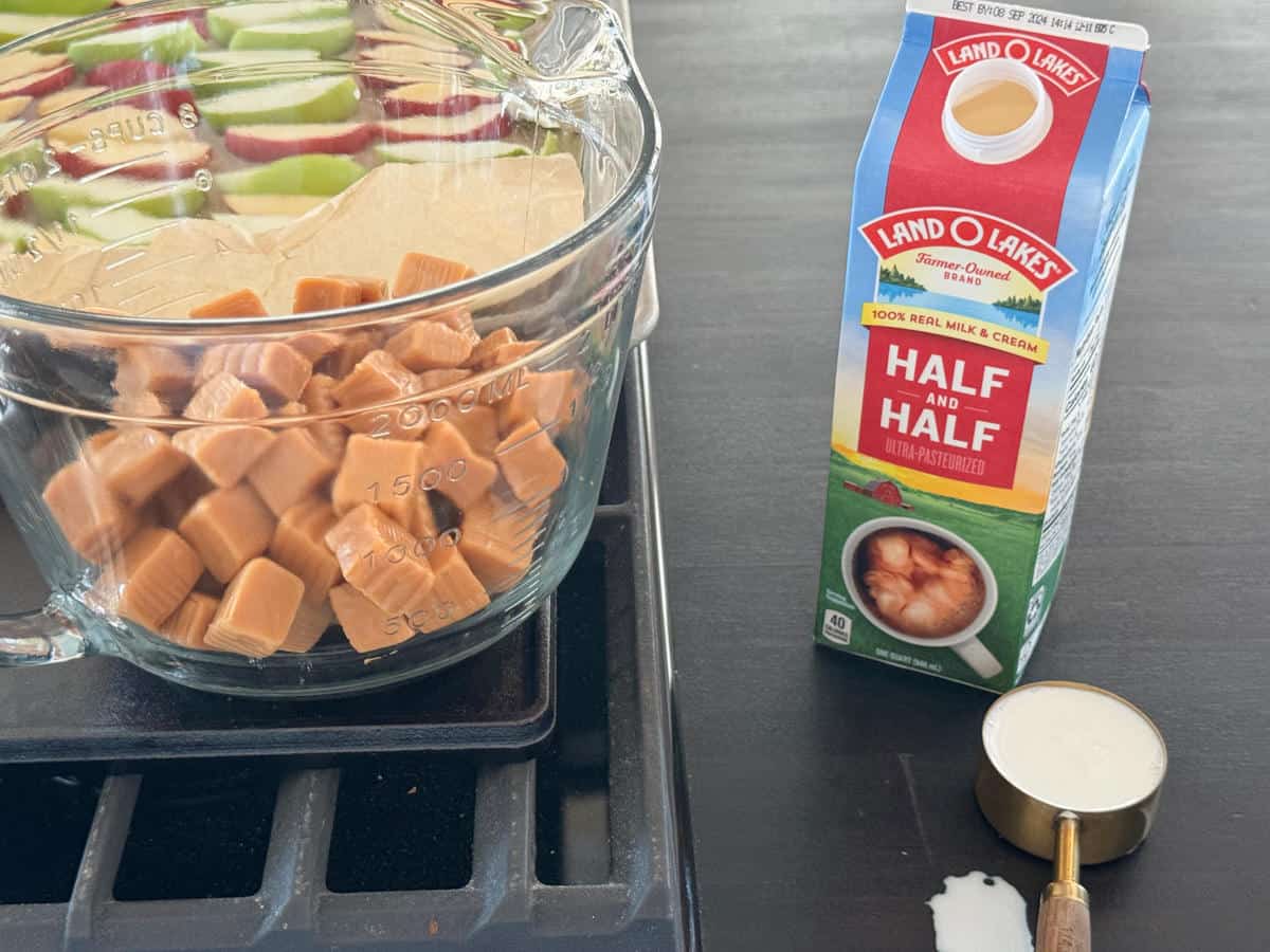 A glass measuring cup filled with caramel squares is placed on a stove. Beside it is a carton of Land O' Lakes half and half cream. A metal measuring cup with some spilled liquid is on the right side, on the stovetop surface.