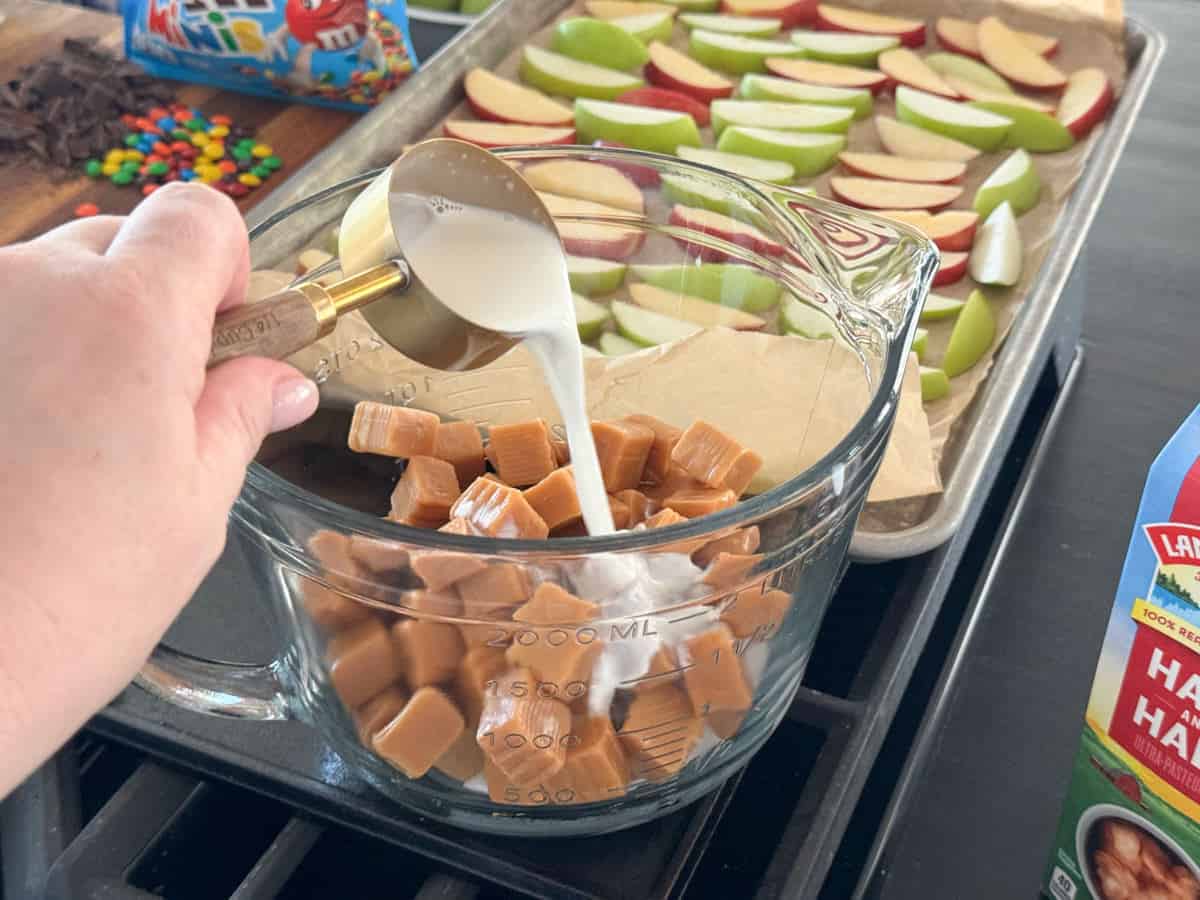 A hand pours milk into a large glass measuring cup filled with caramel candies. In the background, a baking sheet displays sliced green and red apples. Various toppings such as mini chocolates and candy-coated chocolates are positioned nearby.