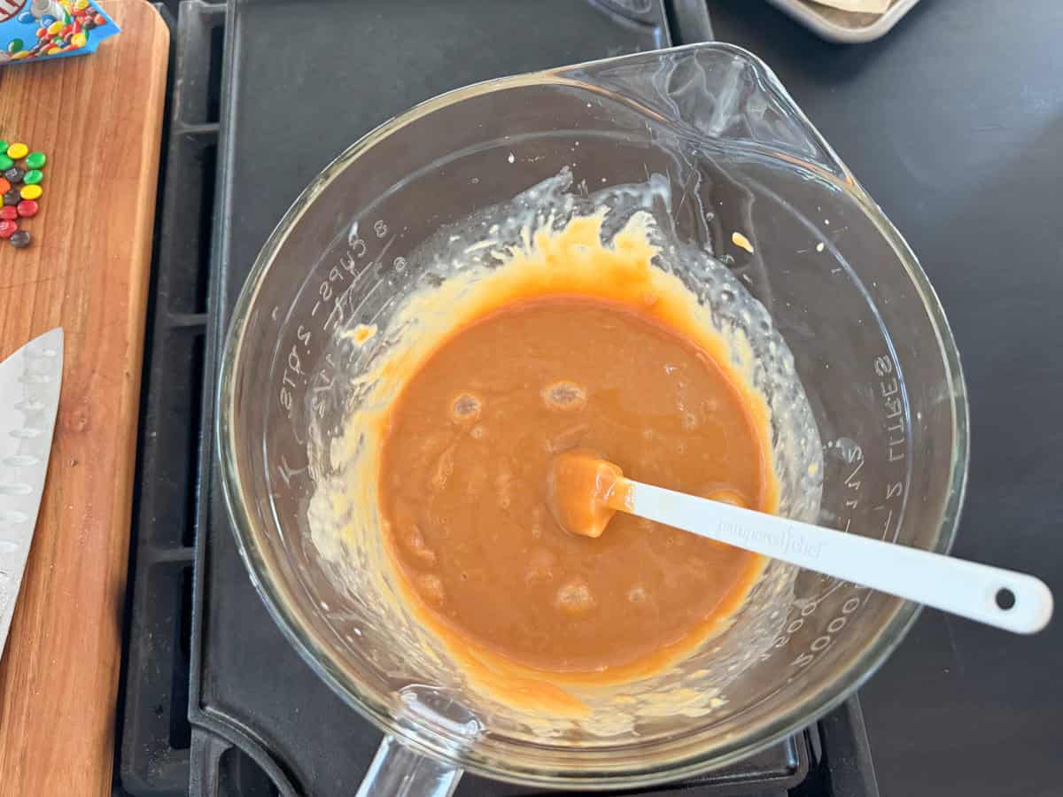 A glass mixing bowl containing a thick, orange-brown mixture with some visible lumps. A white spatula with writing on the handle is resting inside the bowl. The bowl is placed on a dark surface next to a cutting board with a knife and some colorful candy pieces.