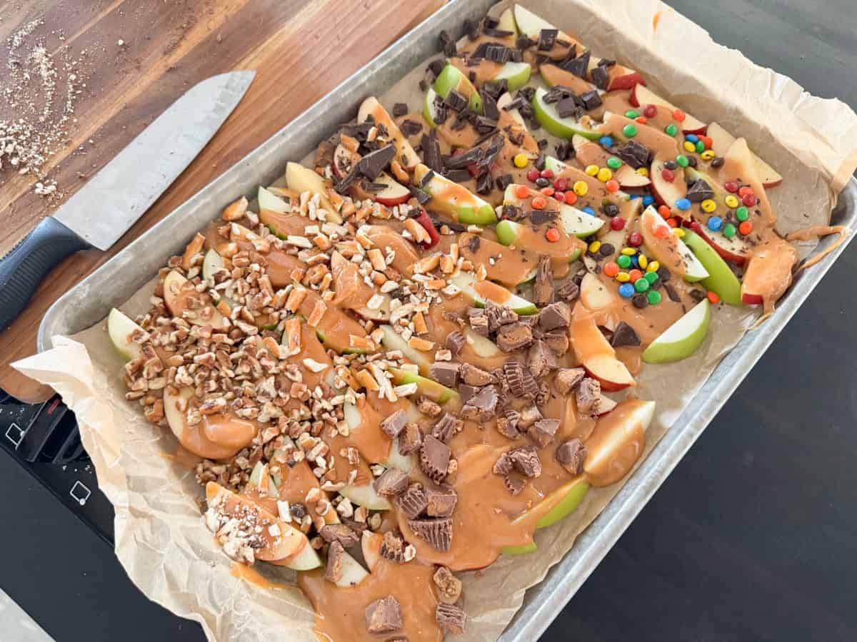 A baking tray lined with parchment paper holds sliced apples topped with various ingredients: chopped nuts, caramel sauce, chocolate pieces, crumbled peanut butter cups, and colorful candy-coated chocolates. A knife rests on the wooden cutting board beside the tray.