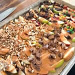 A baking sheet filled with sliced apples topped with different ingredients, including drizzled caramel, chopped nuts, mini candy-coated chocolates, pieces of chocolate, and bits of candy bars. A knife is partially visible on the top left corner.