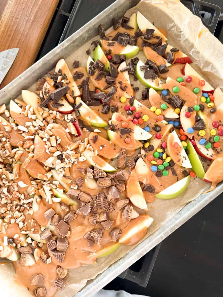 A tray lined with parchment paper holds apple slices covered in various toppings. The toppings include a caramel-like sauce, crushed nuts, chocolate chunks, and colorful candies. The apples are arranged in separate sections based on the toppings.