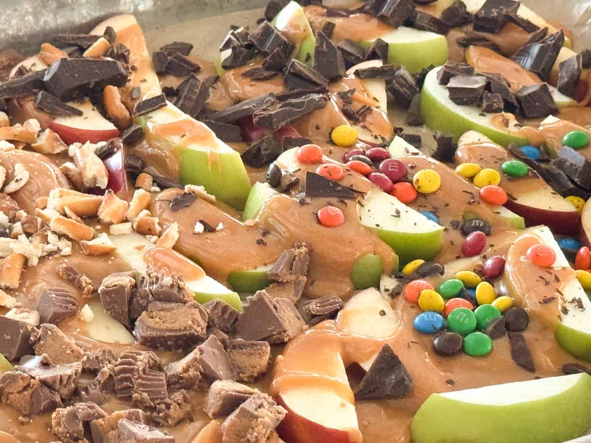 A close-up photo of sliced apples topped with various toppings. The toppings include caramel sauce, chocolate chunks, crushed nuts, mini peanut butter cups, and colorful candy-coated chocolates. The apple slices are arranged side-by-side on a tray.