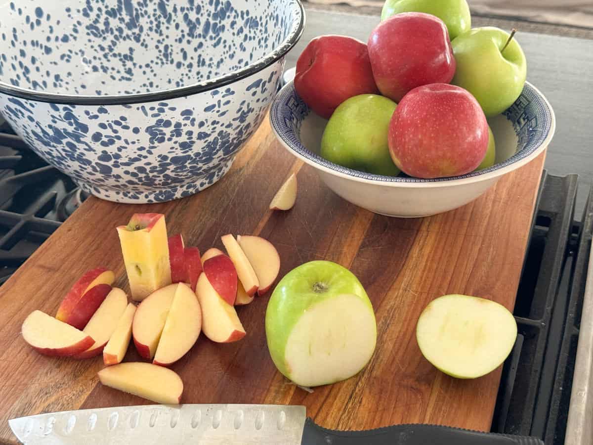A wooden cutting board with sliced apples, a knife, and two bowls in the background. One bowl contains whole red and green apples, while the other is an empty speckled blue and white ceramic bowl.