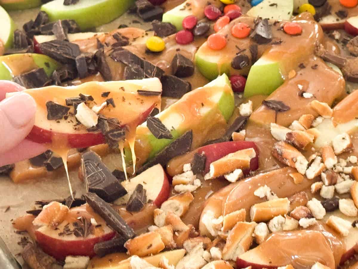 A close-up of caramel apple slices topped with various ingredients including chocolate chunks, pretzel pieces, M&M candies, and nuts. A hand is holding one of the apple slices covered in caramel and chocolate.
