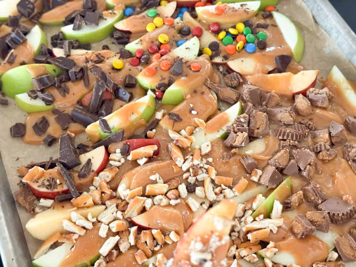 Apple slices on a tray are topped with various toppings, including caramel, chocolate chunks, crushed pretzels, chopped peanuts, and colorful candy-coated chocolates. The tray is lined with parchment paper, and the ingredients are evenly distributed.