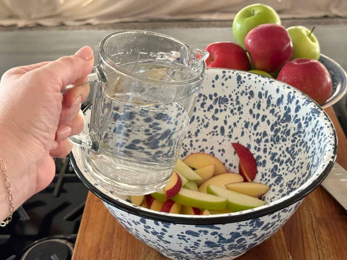 A hand is holding a clear glass jug filled with water, pouring it into a blue-speckled white enamel bowl containing sliced red and green apples. In the background, there are whole apples in a bowl.