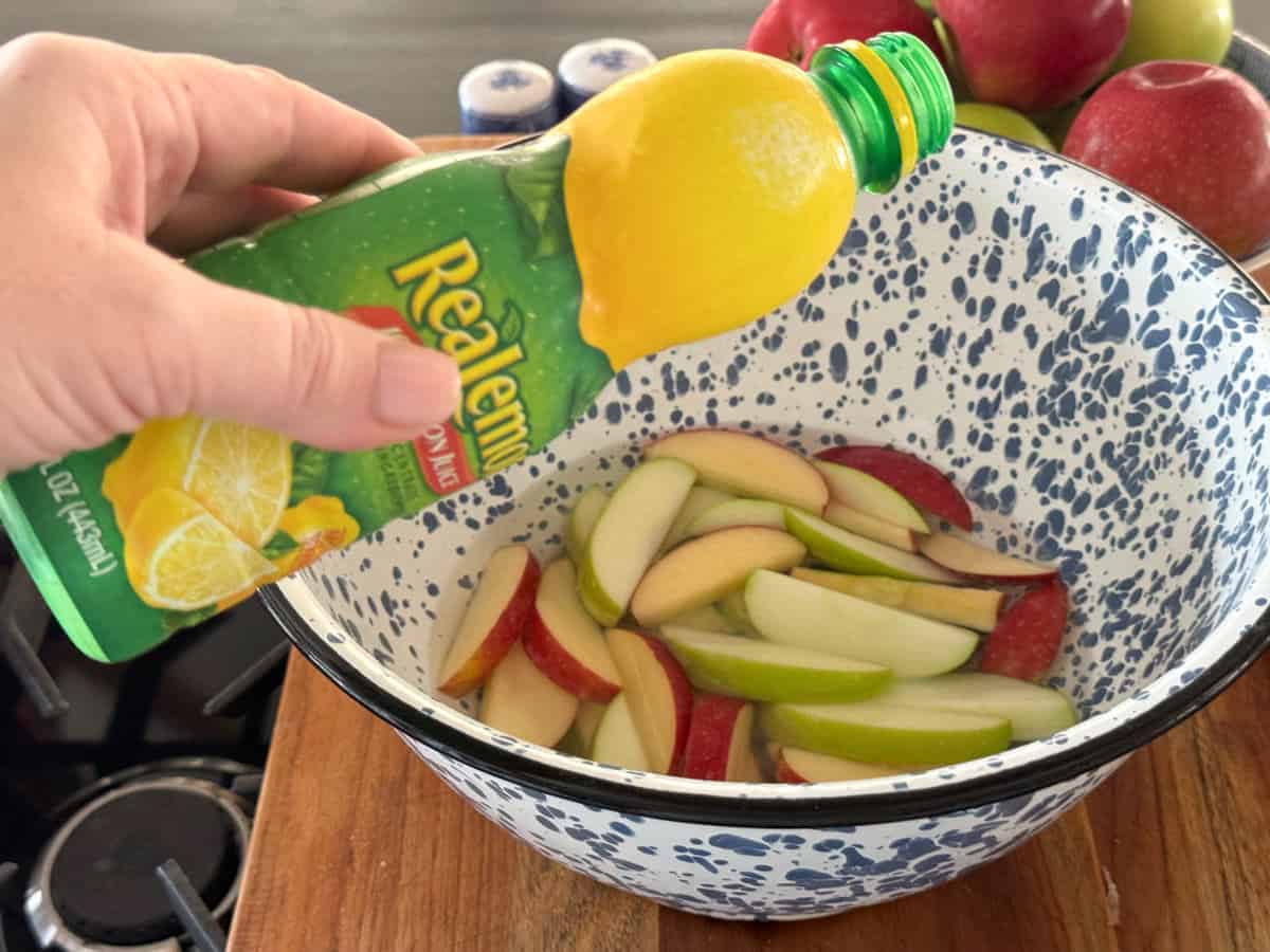 A hand is squeezing a green plastic bottle of ReaLemon lemon juice over a white speckled bowl filled with sliced red and green apples on a wooden surface. There are whole apples and spice containers in the background.