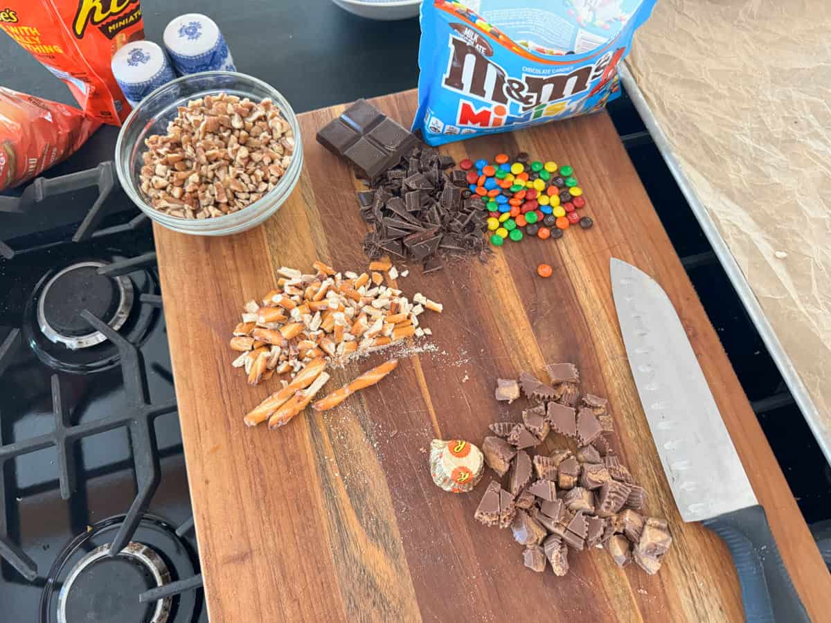 Ingredients on a wooden cutting board are being prepped for baking or cooking. They include chopped pecans, pretzels, dark chocolate, M&M's, and Reese's Peanut Butter Cups. A sharp kitchen knife is also on the board. The background shows a stovetop.