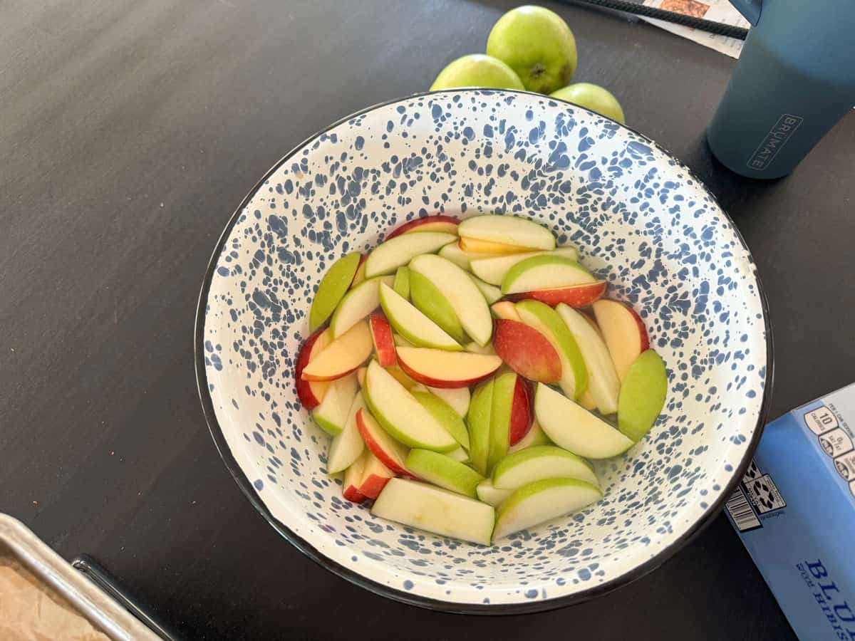 A white and blue speckled bowl on a black table holds slices of red and green apples soaking in water. Three green apples, a blue insulated bottle, and a magazine are placed nearby.