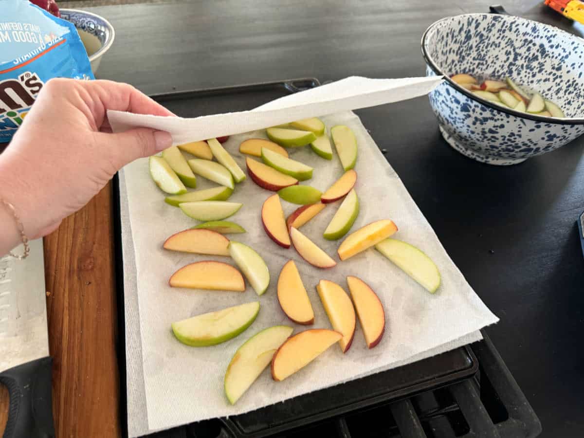 A hand lifts a paper towel over a tray of sliced apples, both red and green, arranged on white paper towels. In the background, there is a blue and white bowl with more apple slices in water. A bag of candy is partially visible to the left.