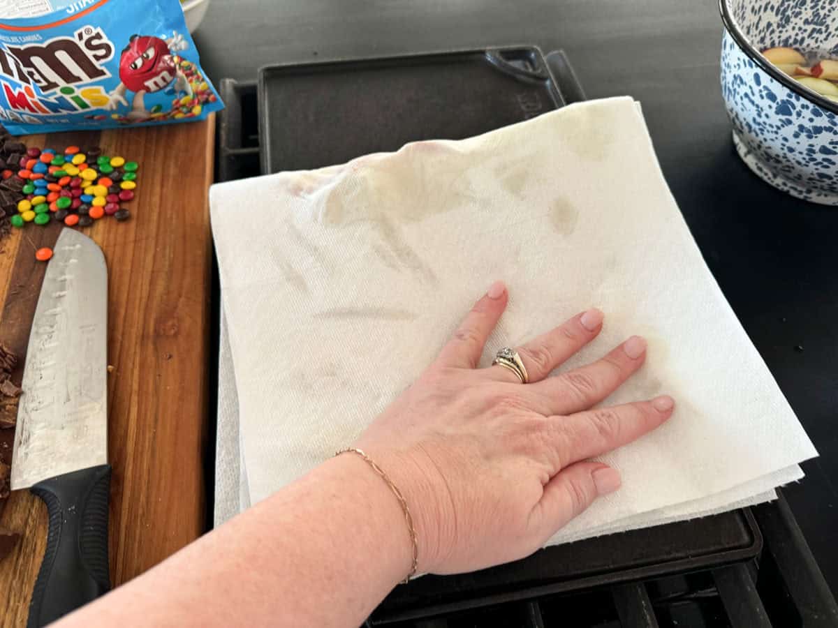 A hand pressing down on a paper towel placed over a griddle. Nearby are a knife with some chocolate residue, an open bag of M&M's, and a bowl containing some mixture. The scene suggests preparation of a sweet dish.