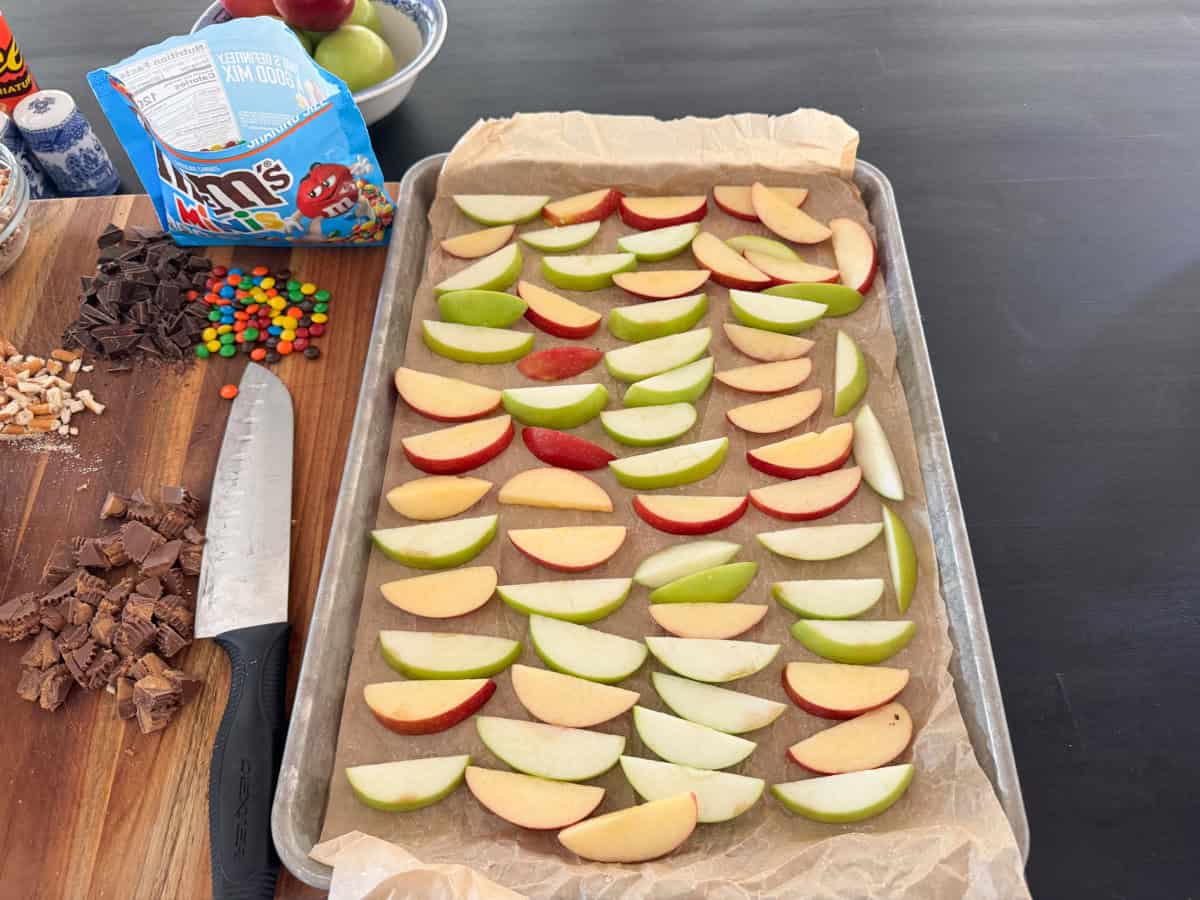 A baking sheet lined with parchment paper displays neatly arranged slices of green and red apples. A large cutting knife rests on the left. Nearby are small bowls containing desserts, including chocolate pieces and colorful candies, with a bag of M&M’s in view.