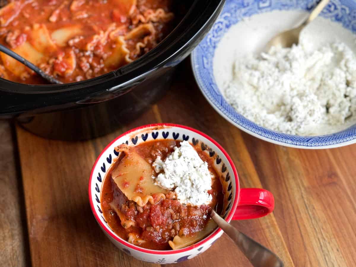 A bowl of lasagna soup topped with a dollop of ricotta cheese sits in a red and white mug on a wooden surface. Nearby, a crockpot filled with lasagna soup and a blue and white bowl containing ricotta cheese with a spoon are visible.