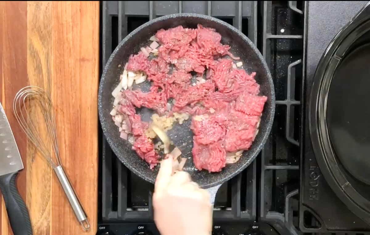 A person is cooking ground beef and chopped onions in a frying pan on a stovetop. A spatula is being used to stir the ingredients. On the wooden counter beside the stove, there is a whisk and a knife with a black handle.