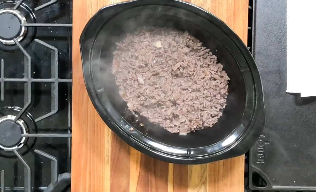 A slow cooker filled with cooked ground meat sits on a wooden cutting board. The slow cooker is positioned next to a stovetop range with burners visible on the left side of the image. Steam is rising from the meat, indicating it is hot.