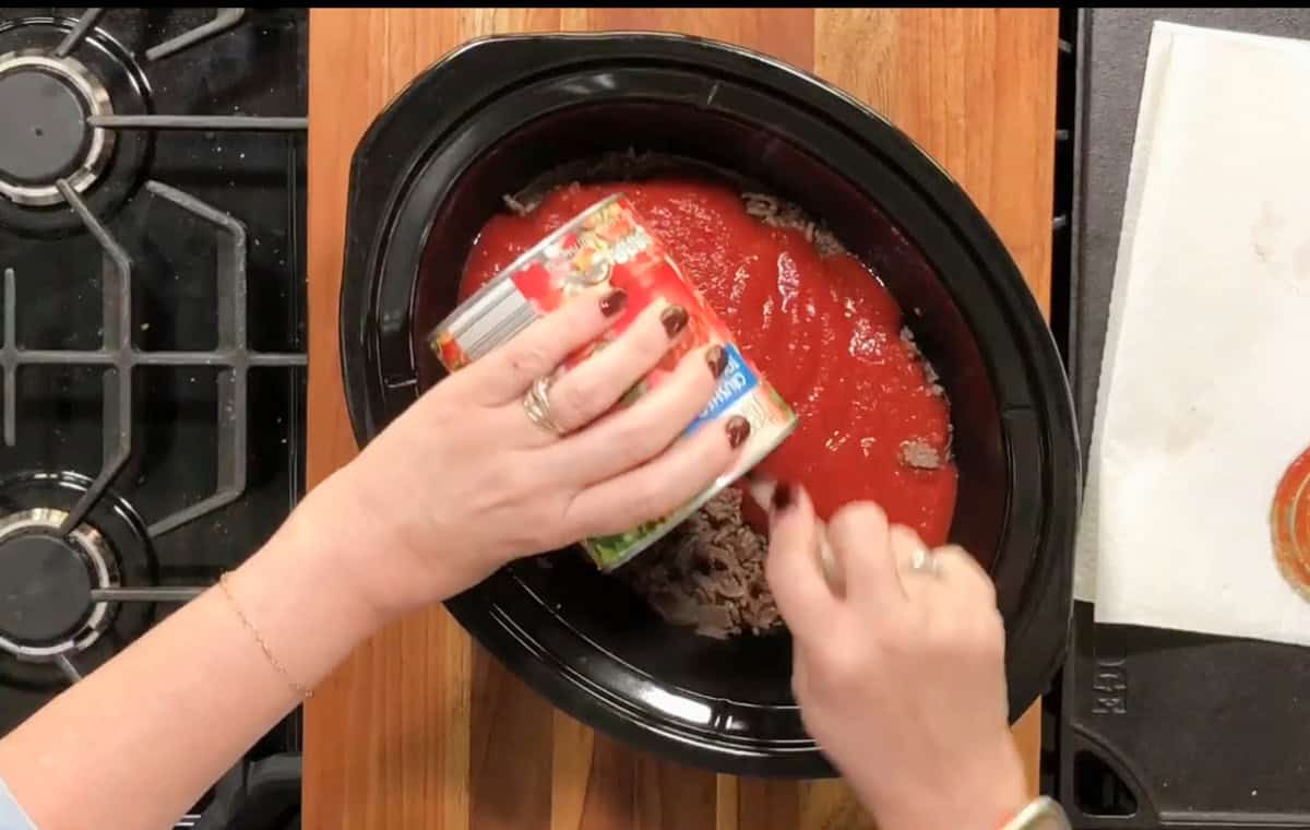A person is seen pouring tomato sauce from a can into a black slow cooker that contains pieces of meat. The cooker is placed on a wooden countertop. A stove is visible on the left side of the image, and a white towel is partially seen on the right.