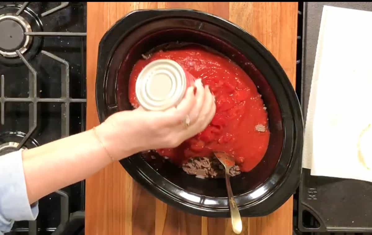 A person's hand is shown pouring tomato sauce from a can into a slow cooker. The slow cooker contains ground beef and is placed on a wooden countertop next to a stove. There is a metal spoon in the slow cooker and a white napkin beside it.